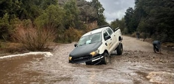 Una camioneta blanca y negra atascada en un río de agua marrón que atraviesa un camino de tierra junto a árboles y rocas