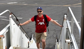 Un hombre con gorra, gafas de sol y camiseta roja de fútbol sujeto a las barandillas de una escalerilla de avión sobre la pista de asfalto
