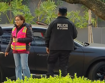 Un policía de espaldas con la frase Policía de la Ciudad y una mujer con chaleco rosa se encuentran junto a un automóvil negro