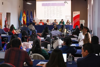 Vista de una sala de conferencias con un panel de seis mujeres en el escenario y una audiencia sentada en mesas azules, con banderas y una pantalla grande al fondo