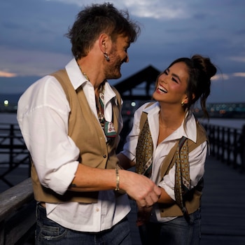 Un hombre y una mujer sonrientes se miran en un muelle de madera. Visten camisa blanca, chaleco marrón, pantalón vaquero. De fondo, agua y luces urbanas