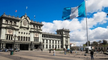 Vista frontal del Palacio Nacional de la Cultura en Guatemala con su fachada gris, una bandera guatemalteca grande ondeando a la derecha y personas en la plaza empedrada