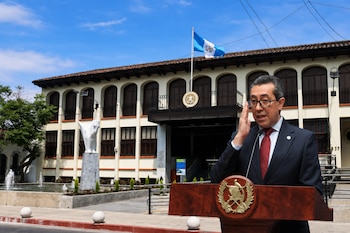 Un hombre con traje, anteojos y corbata habla tras un podio con el escudo de Guatemala ante un edificio oficial.