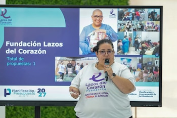 Una mujer de cabello rizado y gafas habla por un micrófono delante de una pantalla gigante que muestra el logo de la Fundación Lazos del Corazón y texto