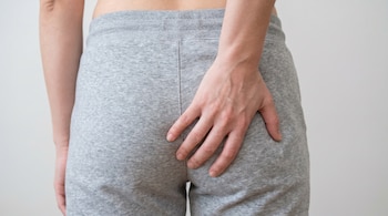 Plano detalle trasero de una persona con pantalón de jogging gris, tocándose la zona de la cola con la mano derecha, sobre fondo blanco.