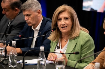 Mujer de cabello rubio, vestida con chaqueta verde, hablando en un micrófono. Botellas de agua y hombres sentados al fondo