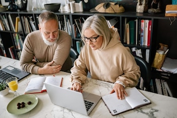 Hombre barbudo y mujer con gafas en una mesa de mármol, trabajando en laptops y cuadernos abiertos. Un vaso de jugo y bombones en un plato. Estanterías con libros detrás