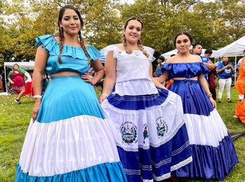 Tres mujeres. Visten trajes folclóricos azul y blanco con faldas largas y blusas cortas. La falda del centro lleva el escudo de El Salvador. Fondo de personas y árboles