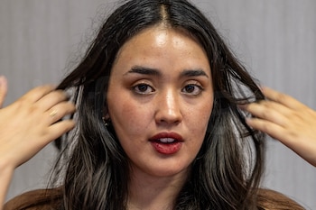Un primer plano de la cantante y escritora peruana Elisa Tokeshi mirando fijamente a la cámara mientras se acomoda el cabello con ambas manos sobre un fondo gris neutro, capturando su espontaneidad característica de las redes sociales.
