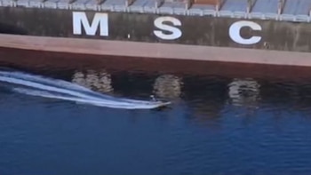 Vista lateral de un gran buque de carga con las letras 'MSC' en su casco, mientras una pequeña lancha a motor deja una estela sobre el agua oscura