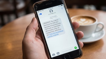 Mano de persona sujeta un teléfono móvil con pantalla agrietada que muestra un mensaje de texto de "ALERTABANCO" sobre una cuenta bancaria bloqueada.