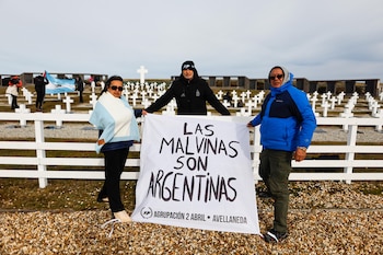 Tres personas despliegan una pancarta blanca frente a hileras de cruces blancas en un cementerio