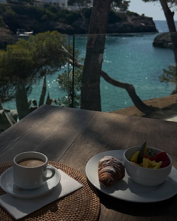 Una mesa de madera con un café con leche, un croissant espolvoreado con azúcar y un tazón con fruta cortada. Al fondo se observan el mar, acantilados y vegetación