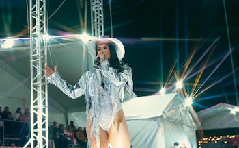 Mujer con sombrero vaquero, atuendo plateado con flecos, y mallas de red cantando en un micrófono sobre un escenario iluminado por luces
