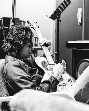 Fotografía en blanco y negro de un hombre de cabello rizado sentado en un sillón que toca una guitarra eléctrica en un estudio