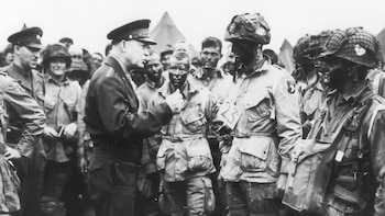 Fotografía en blanco y negro de Dwight D. Eisenhower uniformado frente a un grupo de soldados con cascos, equipo militar y rostros pintados