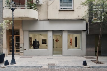 Interior de una galería de arte con paredes blancas y suelo gris, mostrando varias obras: una casa negra en patas, un cuadro texturizado, dos lienzos gráficos, y una escultura suspendida