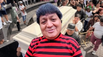Hombre, Pompinchú, sonríe con polo rojo y negro. Detrás, un ataúd blanco es llevado por personas en una escena borrosa de procesión fúnebre.