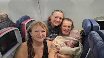 Tres mujeres sonríen a la cámara dentro de la cabina de un avión; una de ellas sostiene a un bebé recién nacido envuelto en una manta clara