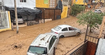 Calle de tierra inundada de barro, dos coches plateados semisumergidos, casas coloridas en los laterales y un poste de luz al centro