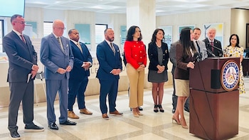 Una mujer habla en un podio con el sello del Senado de Florida, rodeada por ocho personas en un salón de eventos