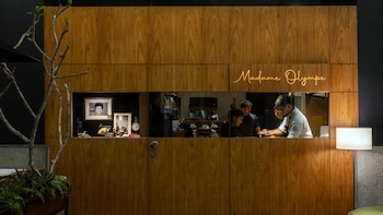 Pared de madera con letrero de neón 'Madame Olympe'. Una ventana muestra chefs en la cocina. A la izquierda, un nicho con fotos y objetos; a la derecha, una lámpara