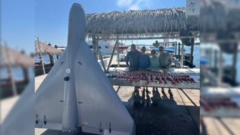 Un dron gris en primer plano y cuatro personas detrás de una mesa con peces en un muelle marítimo