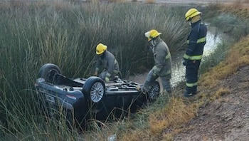 Tres bomberos con cascos amarillos y uniformes oscuros observan un SUV negro volcado de lado en una zanja con agua y vegetación alta
