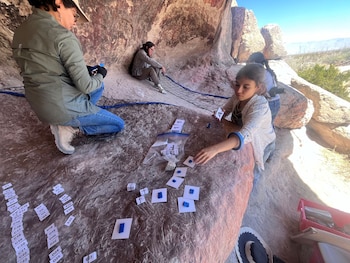 Tres personas trabajan dentro de un abrigo rocoso, clasificando pequeños fragmentos de roca con etiquetas en la superficie de una roca y en el suelo