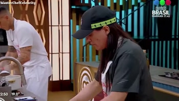 Un hombre con gorra negra y delantal blanco prepara alimentos en una cocina de televisión junto a otro participante al fondo