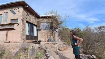 Una mujer frente a una vivienda de adobe y madera con ventanas recicladas sobre un terreno rocoso en la sierra bajo un cielo despejado