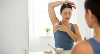 Mujer joven aplica polvo blanco en su axila izquierda con la mano derecha, mirándose en un espejo en un baño. Un cepillo de dientes y jabón están en la encimera.