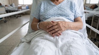 Manos de una persona cruzadas sobre una sábana blanca en una cama de hospital, con una bata estampada y un brazalete de identificación.