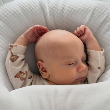 Primer plano de un bebé durmiendo de lado en una cuna blanca, con sus brazos levantados y manos cerca de la cabeza. Viste un pijama claro con ositos