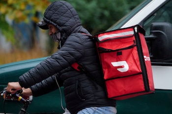 Una persona con chaqueta acolchada negra, gorra y mascarilla, pedalea en bicicleta llevando una mochila de entrega DoorDash roja en su espalda