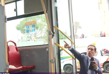 Reportero con micrófono dentro de un bus con asientos rojos y barras amarillas. Por la ventana se aprecian ambulancias y personas