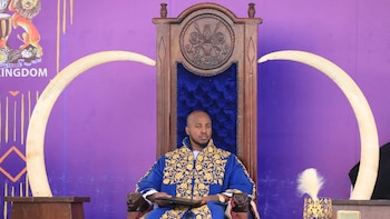 Hombre con atuendo azul y dorado sentado en un trono de madera flanqueado por dos colmillos de elefante frente a un fondo morado