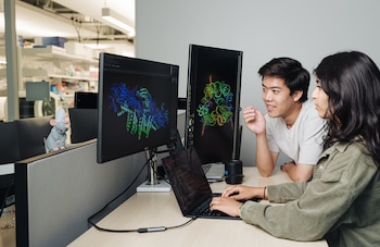 Dos personas frente a tres computadoras, con monitores que muestran diagramas moleculares. Un peluche de rata chef adorna el escritorio en un fondo de laboratorio