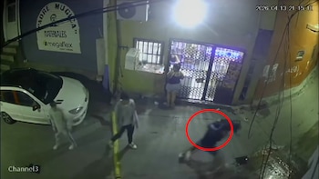 Vista aérea de cámara de seguridad nocturna mostrando una persona cayendo al suelo (circulada en rojo), con otras figuras, un auto blanco y edificios alrededor