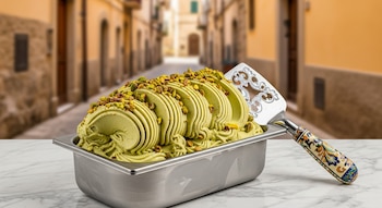 Gelato de pistacho verde brillante en un contenedor metálico sobre un mostrador de mármol, con una pala decorativa y un pueblo italiano borroso detrás.