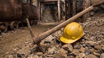 Pica de madera y metal oxidado, casco de minero amarillo sobre rocas y tierra, dos vagonetas oxidadas sobre rieles, túnel de mina con soporte de madera.