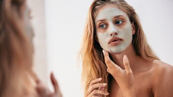 Woman looking in the mirror with mask on her face. Female applying facial cosmetic mask in bathroom.