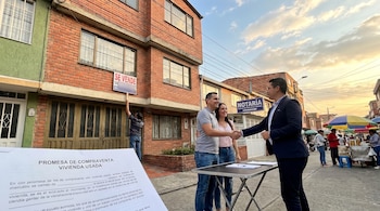 Una pareja sonriente estrecha la mano de un notario frente a un edificio de ladrillo con un cartel de 'Se Vende'. En primer plano, documentos sobre una mesa.
