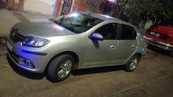 Un sedán plateado estacionado de noche en una calle de cemento. Se observa un reflejo de luz azul en el frente y parte de un vehículo rojo detrás