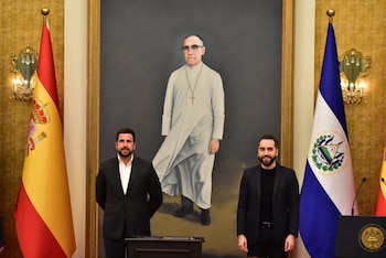 Dos hombres con traje posan frente a una pintura religiosa, flanqueados por la bandera de España y la de El Salvador