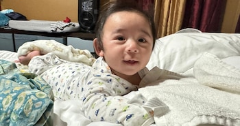 Bebê sorridente com macacão estampado, deitado na cama bagunçada com lençóis e toalhas brancos e verdes