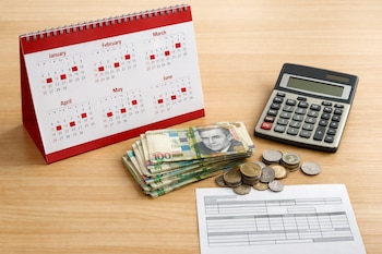 Calendario de escritorio de enero a junio, pila de billetes de cien soles peruanos, monedas, calculadora y un recibo de sueldo genérico sobre una mesa de madera.