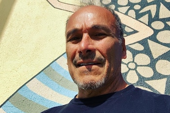 Primer plano de un hombre de mediana edad con cabello grisáceo, bigote y barba corta, usando camiseta oscura, frente a una pared con patrones geométricos