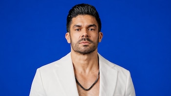 Retrato de un hombre de cabello oscuro peinado hacia atrás con saco blanco y collar sobre un fondo azul uniforme