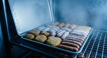 Primer plano de nuggets y salchichas vegetales congelados en una bandeja metálica dentro de un congelador industrial con vapor frío